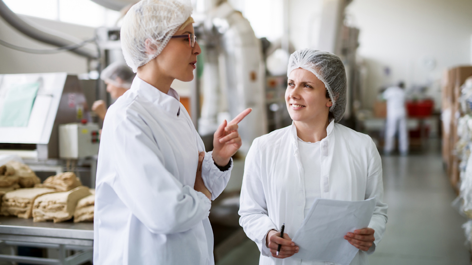 Two female food safety technicians standing and reviewing a report, inside a food manufacturing facility.