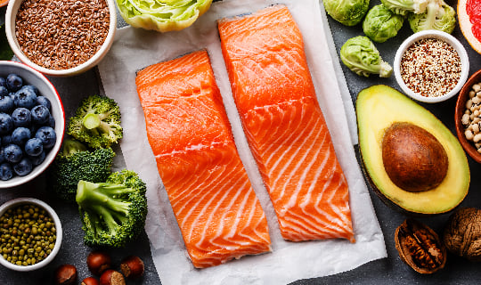 Two salmon states on cutting board with raw avocado, broccoli, blueberries, and spices in bowls around the board.
