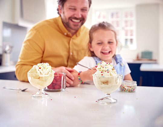 Father And Daughter In Kitchen Decorating Ice Cream Dessert With Cream And Sprinkles