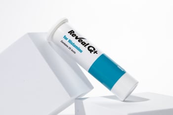Reveal® Q+ for Histamine test kit tube resting diagonally on white geometric display blocks, with teal accent label and “contains 25 tests” visible on packaging.