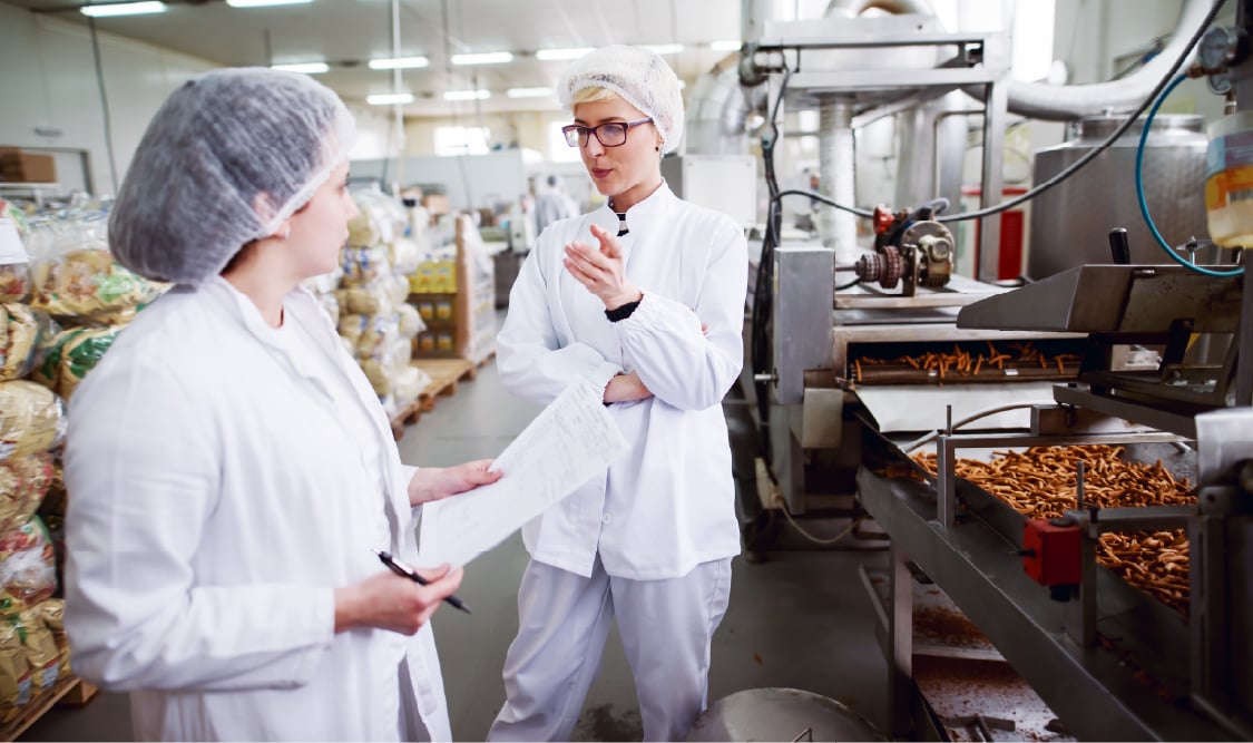 Two female food safety technicians standing and talking inside a food manufacturing facility