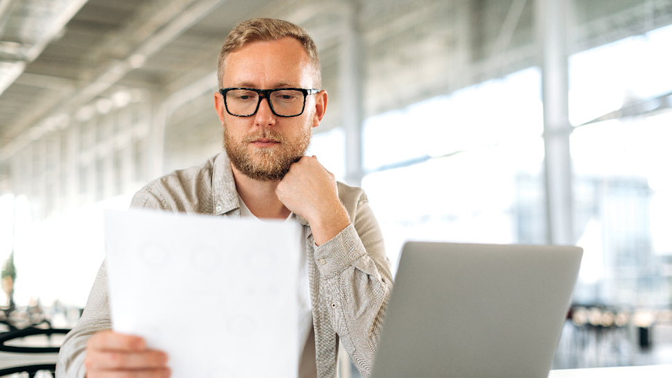 Man reading a sheet of paper next to his lap top.
