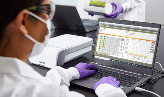 A lab technician in a white coat and purple gloves opens a Neogen molecular detection system for pathogen testing on a laboratory bench.