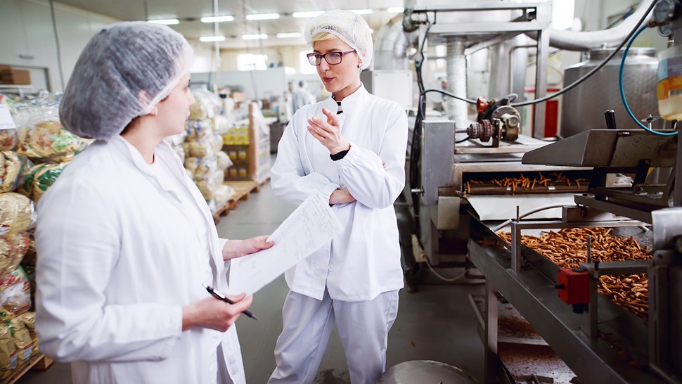 Food safety professionals in protective clothing reviewing documentation beside a production line in a food manufacturing facility.